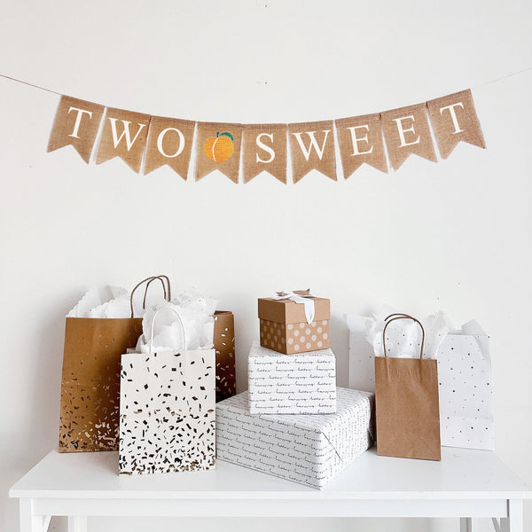 This is a burlap banner made from 4 inch by 6 inch fishtail shaped flags. The words Two Sweet are stenciled in white, one letter per flag. There is an image of a peach on a flag between the two words.