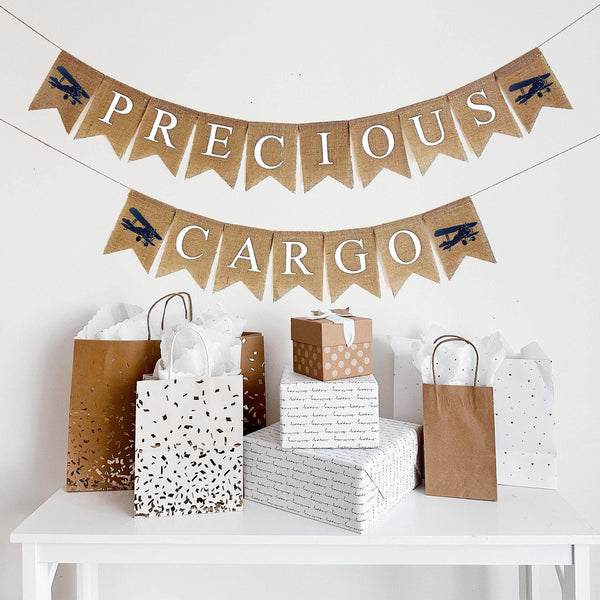 This is a burlap banner made of 4.5 by 6.5 inch fishtail flags. The top string says PRECIOUS spelled in white capital letters with an image of a navy plane on either end. The bottom string says CARGO in white letters with a navy plane on each end.