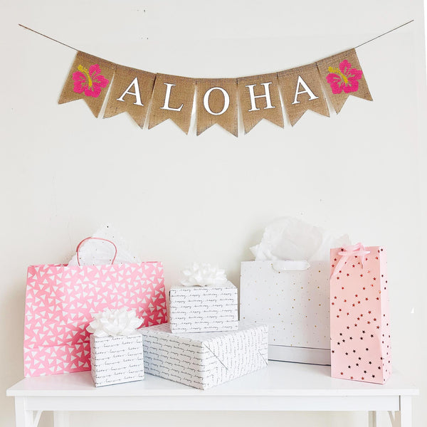 Tropical Aloha Luau Burlap Banner. Aloha is written in white lettering on burlap flags with a hand stenciled hot pink hibiscus flower on each end of the word. The hibiscus is a bright pink base with yellow center.