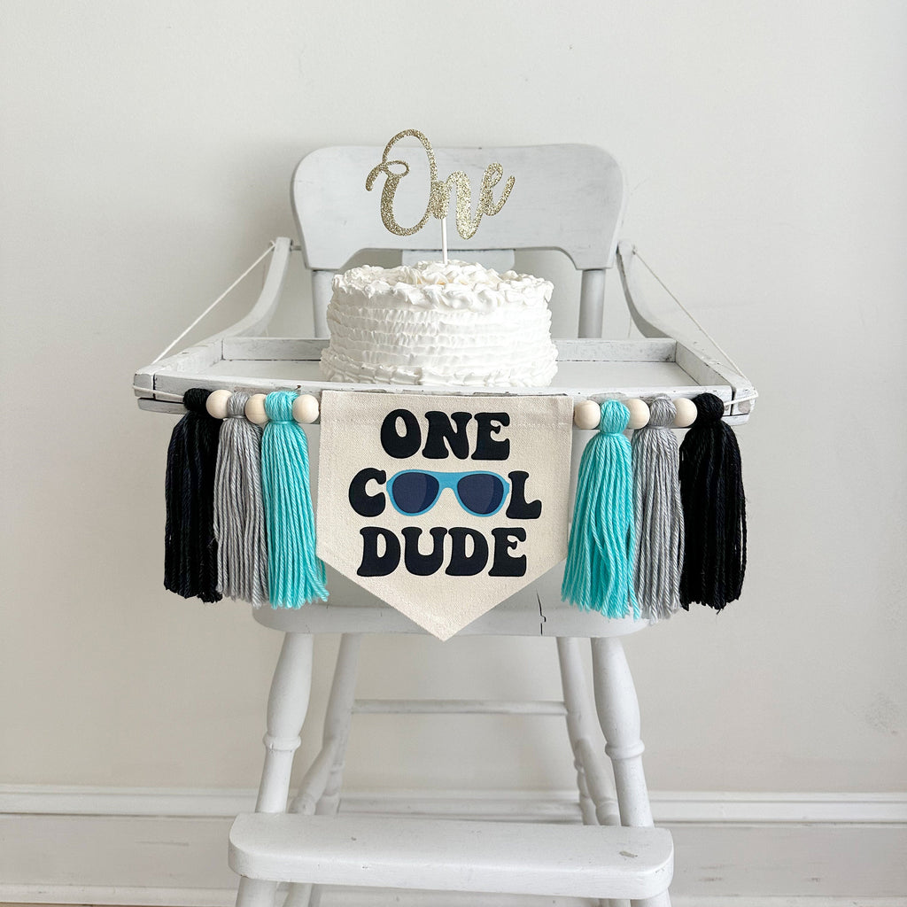This is a 6.5 x 7 inch pennant center flag with the words One Cool Dude in a chunky black retro font. A pair of blue sunglasses make the O&#39;s. There are 3 tassels on either side that are light aqua, gray and black in color with wooden beads in between