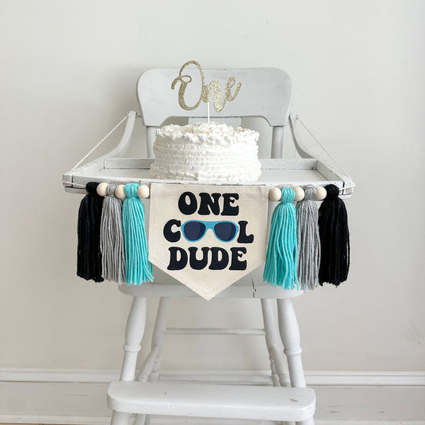This is a 6.5 x 7 inch pennant center flag with the words One Cool Dude in a chunky black retro font. A pair of blue sunglasses make the O&#39;s. There are 3 tassels on either side that are light aqua, gray and black in color with wooden beads in between