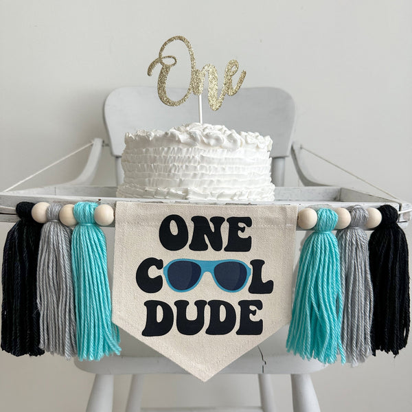 This is a 6.5 x 7 inch center flag with the words One Cool Dude in a chunky black retro font. A pair of blue sunglasses make the O&#39;s. There are 3 tassels on either side that are light aqua, gray and black in color with wooden beads in between.