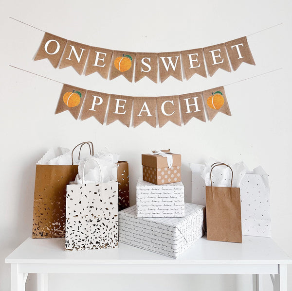 This is a burlap banner on two strings made of fishtail shaped flags with one letter on each flag. The top line says One Sweet in white letters with a peach in between. The bottom line says Peach in white letters with a peach on either end.