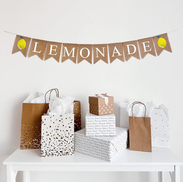 This is a burlap banner made of individual 4.5 by 6.5 inch fishtail shaped flags. The word LEMONADE is spelled out in white letters. There is an image of a lemon on either end of the word LEMONADE.