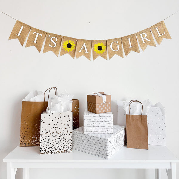 this baby shower banner reads It&#39;s A Girl in white lettering with a sunflower image flag in between each word. The sunflowers are hand stenciled with a vibrant yellow petals with a dark brown center.
