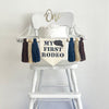This is a banner that can hang on a highchair with a canvas center flag with the words MY FIRST RODEO in navy letters with a cowboy hat at the top. There is a navy, tan and brown yarn tassel hanging down on either side of the center banner.
