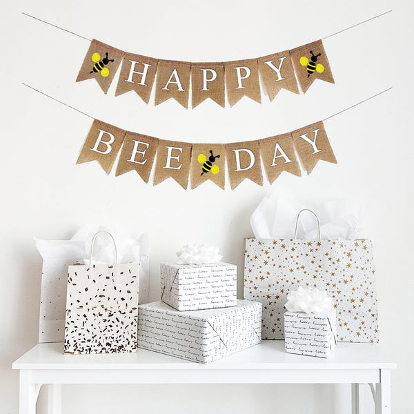 A two tiered burlap banner. The first tier reads HAPPY with a yellow bee icon on either side of the word. The second tier reads BEE DAY with a yellow bee icon between the words.
