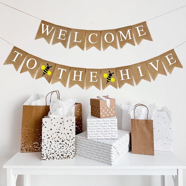 A two tiered burlap banner. The first tier reads welcome. The second tier reads to the hive with a yellow and black bumblee icon between the words.