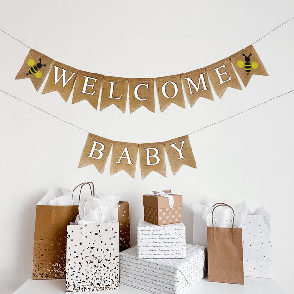 A two tiered burlap banner. The first tier reads welcome with a yellow and black bumblebee icon on each side of the word. The second tier reads baby.