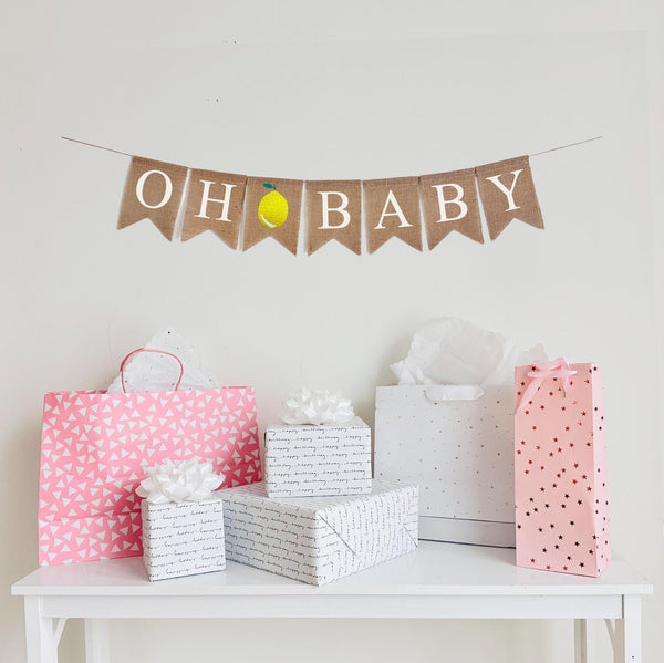 This is a burlap banner made of 4.5 inch by 6.5 inch fishtail shaped flags. It is stenciled with the words OH BABY in white letters with one letter on each flag. There is an image of a lemon with a small green leaf between the two words.