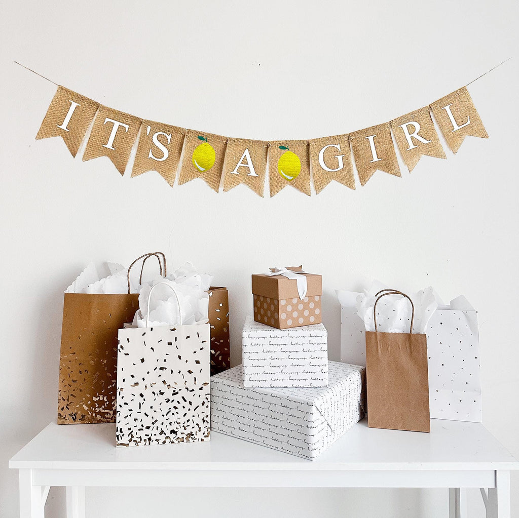 This is a burlap banner made of individual fishtail shaped flags that measure 4.5 by 6.5 inches each. Each flag is stenciled with a white letter. The banner says IT&#39;S A GIRL with an image of a yellow lemon in between each word.