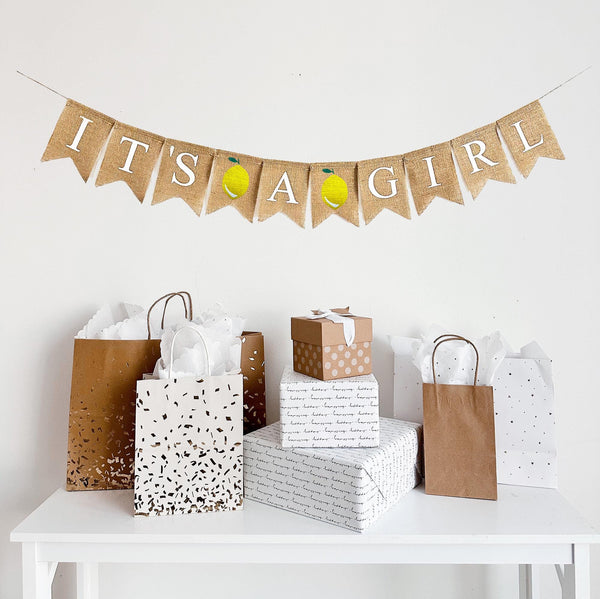This is a burlap banner made of individual fishtail shaped flags that measure 4.5 by 6.5 inches each. Each flag is stenciled with a white letter. The banner says IT&#39;S A GIRL with an image of a yellow lemon in between each word.