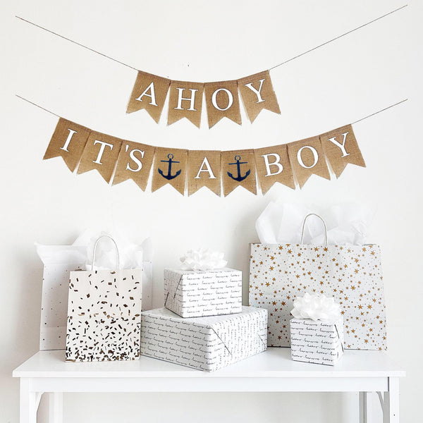 This is a burlap made from 4.5 by 6.5 inch individual fishtail shaped flags. The top string says AHOY in white letters. The bottom string says IT&#39;S A BOY in white letters. There is an image of a navy blue anchor in between the words on the bottom.
