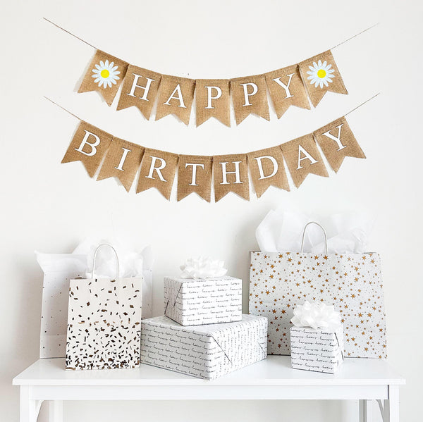 A happy birthday burlap banner strung on two strings. The happy is at the top and the birthday below.  Hand painted daisies are at the beginning and end of the word happy.  The daisies are painted in white with a yellow center.