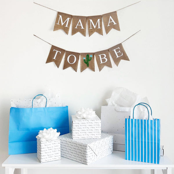 A mama to be burlap banner strung on two strings. The mama is at the top and the to be is below.  Hand painted cacti are in between the words to and be.  The cacti are painted in green with light blue flowers.