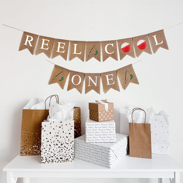 A two tiered burlap banner. The first tier reads reel cool with an image of a fishing rod with a green fish on the line between the words and red and white bobbers for the o&#39;s. The second line reads one with the same fishing rod image on each side.