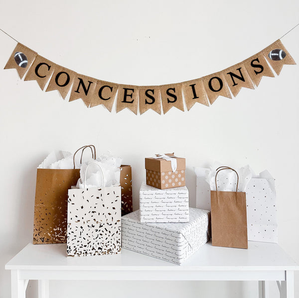 This is a burlap banner made of 4.5 by 6.5 inch fishtail shaped flags. It spells CONCESSIONS in black capital letters and has a hand stenciled image of a football on either end.