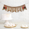 This is a burlap banner made of 4.5 by 6.5 inch fishtail shaped flags. The word ONE is spelled out in white lowercase letters. There is a hand stenciled image of a red tractor on either end.