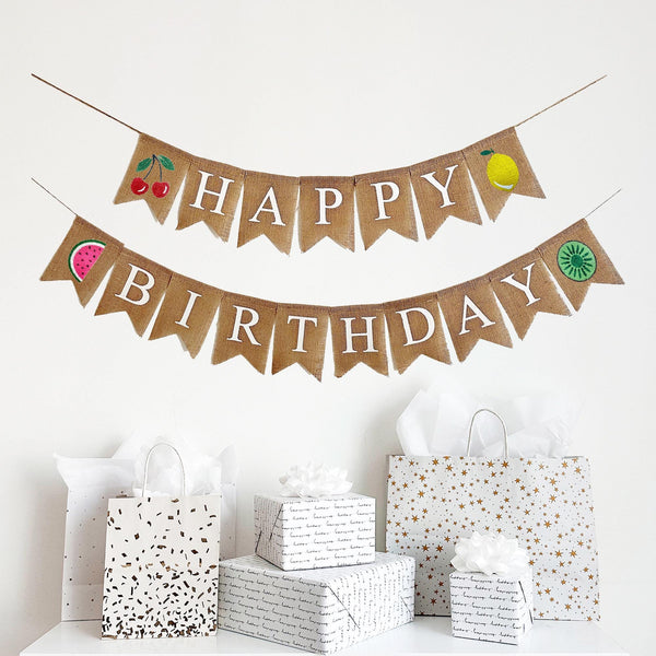 A two tiered burlap banner. The first tier reads happy with a red and green cherry image on one end and a yellow and green lemon on the other end. The second tier reads birthday with a pink watermelon on one end and a green kiwi on the other end.