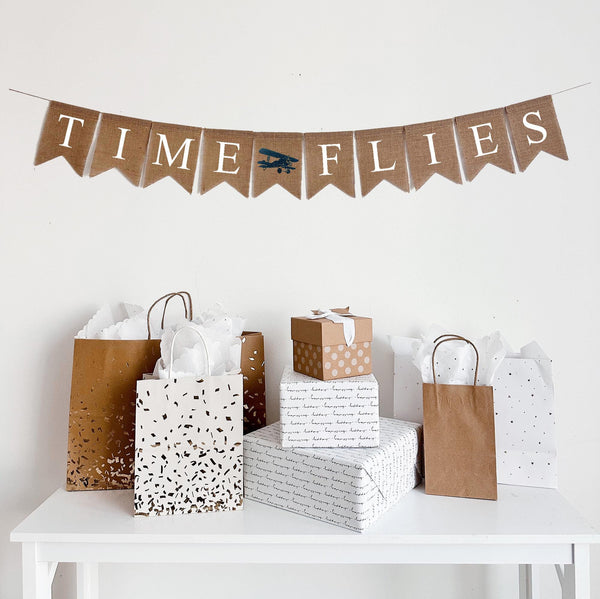 This is a burlap banner made of 4.5 inch by 6.5 inch individual fishtail shaped flags. The words TIME FLIES is stenciled in white capital letters. In between the words, there is an image of a navy vintage biplane.