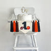 This is a canvas first birthday flag measuring 5x7 inches. It has a large black number 1 with a football, baseball and basketball around it. On each side there is a brown, orange and black yarn tassel, separated by wooden beads.