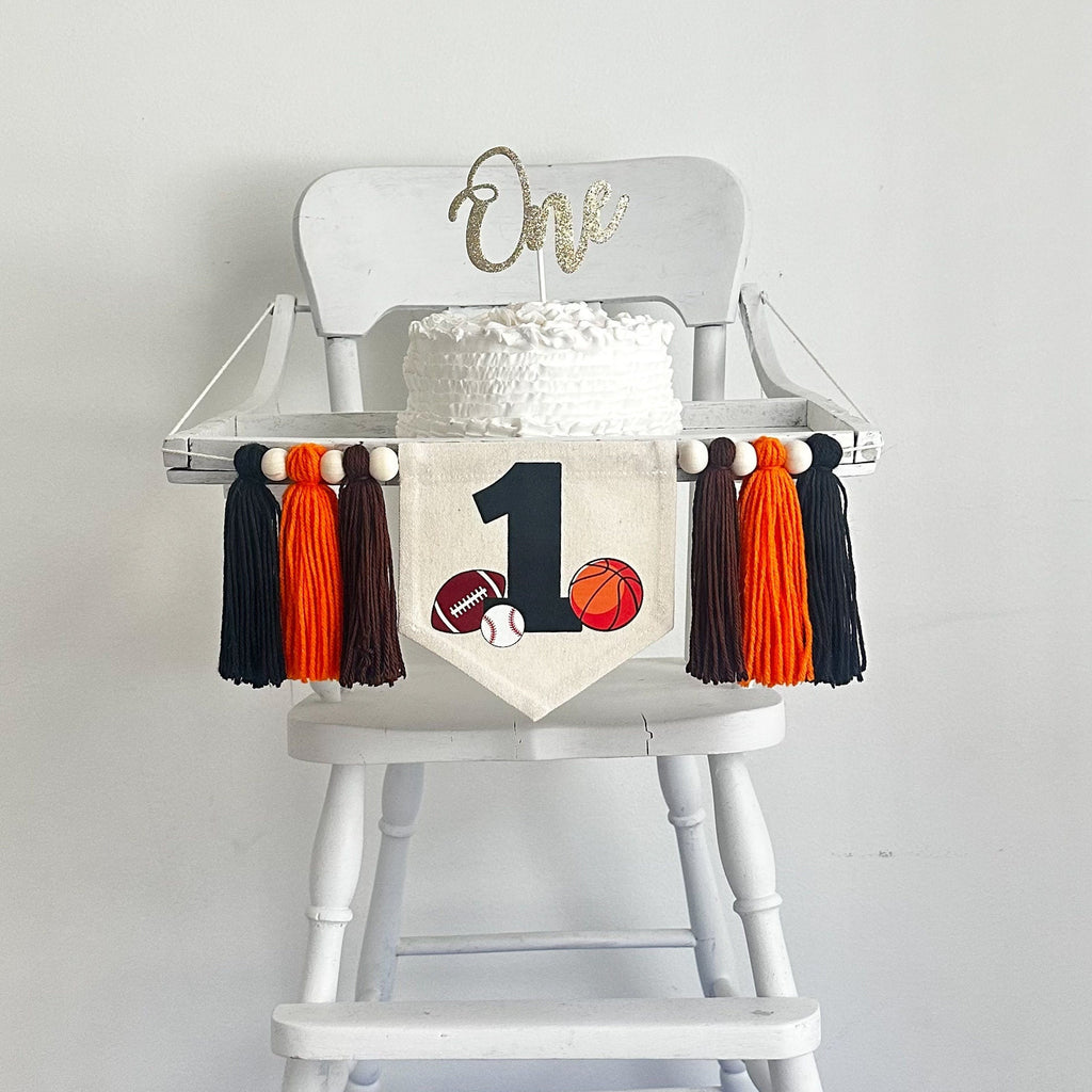 This is a canvas first birthday flag measuring 5x7 inches. It has a large black number 1 with a football, baseball and basketball around it. On each side there is a brown, orange and black yarn tassel, separated by wooden beads.