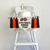 This is a canvas banner with a football, basketball and baseball on it. Below the balls it says the child&#39;s name followed by the words ROOKIE YEAR, all in black. On each side it has a brow, orange and black tassel, each separated by a wooden bead.