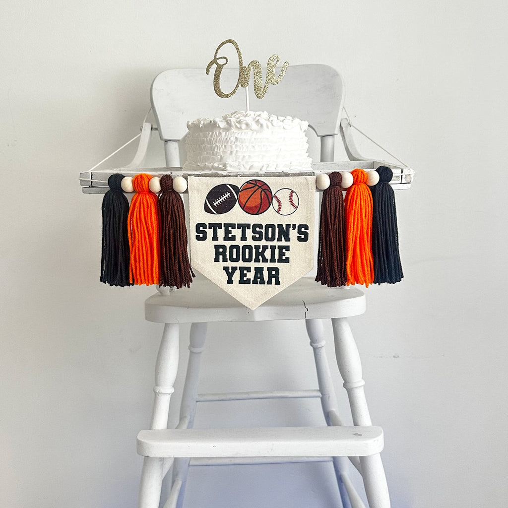 This is a canvas banner with a football, basketball and baseball on it. Below the balls it says the child&#39;s name followed by the words ROOKIE YEAR, all in black. On each side it has a brow, orange and black tassel, each separated by a wooden bead.