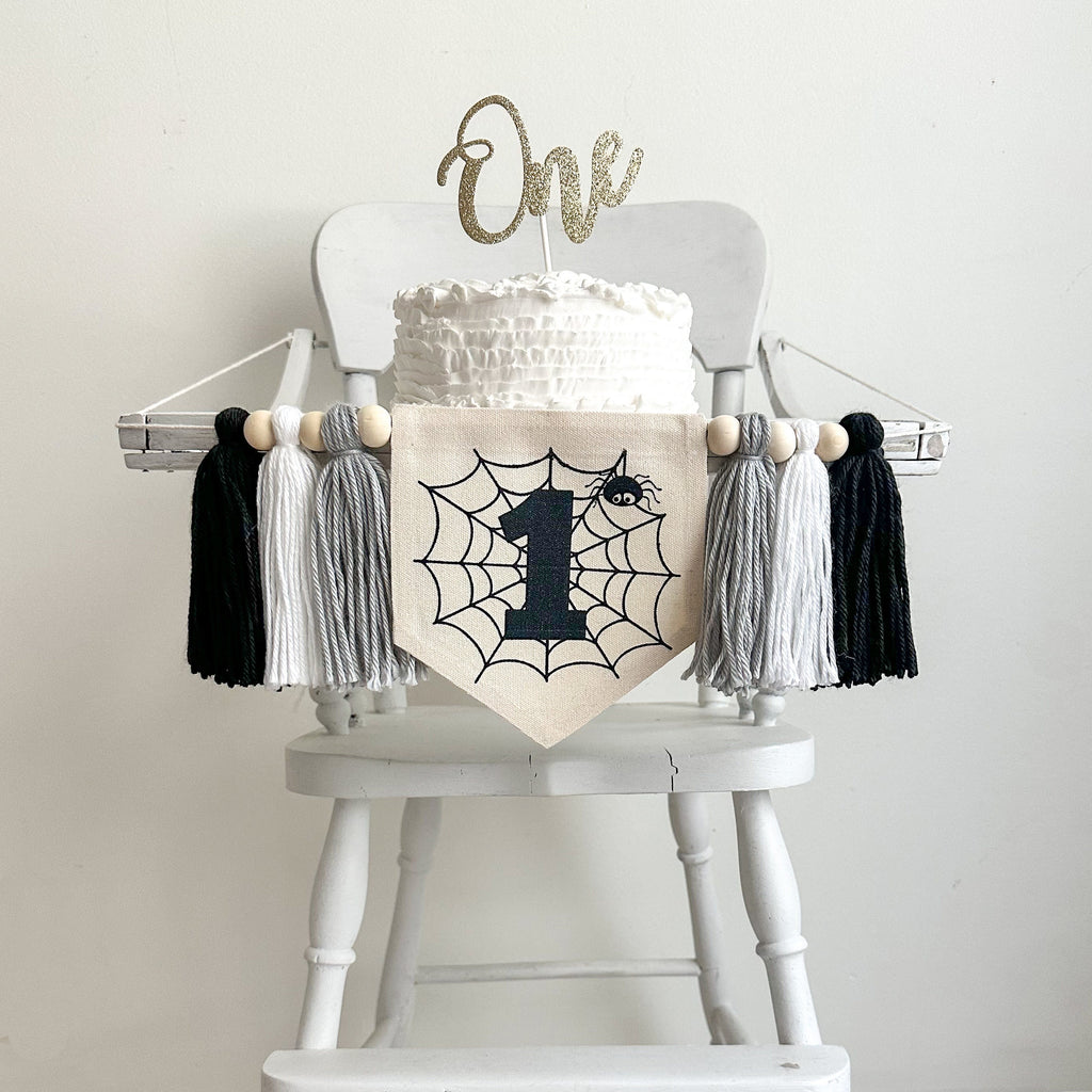 A 5x7 canvas banner with a spider web in the middle with a cute spider in the upper right corner. There is a black number 1 in the middle of the web. There is a gray, white and black tassel on each side of the flag, each separated by a wooden bead.