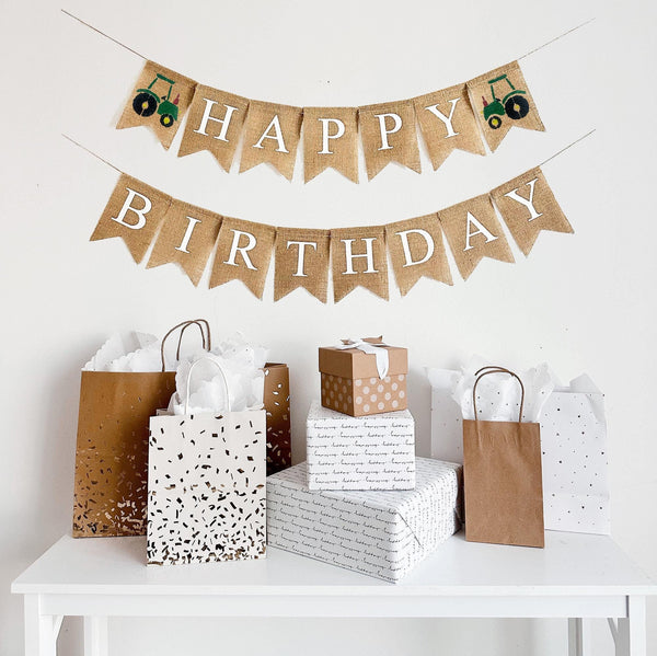 This is a burlap banner made of 4.5 by 6.5 inch fishtail shaped flags. The top string says HAPPY in white capital letters. There is an image of a green tractor on each end of the word. The bottom string says BIRTHDAY in white capital letters.