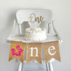 Pink Hibiscus first birthday burlap banner. With a pink flower as the O followed by n e in white letters, this high chair banner is perfect for a tropical first birthday party.
