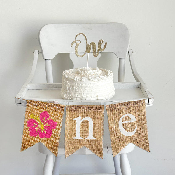 Pink Hibiscus first birthday burlap banner. With a pink flower as the O followed by n e in white letters, this high chair banner is perfect for a tropical first birthday party.