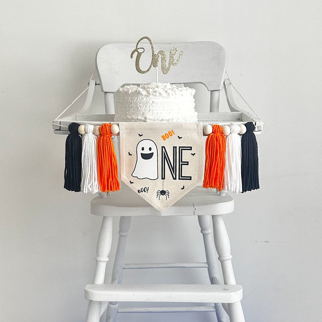 This is a 5x7 inch canvas banner with a ghost image followed by black letters NE to spell the word ONE. It has a spider, bats and a tiny word BOO in orange. There are orange, white and black tassels on each side separated by wooden beads.
