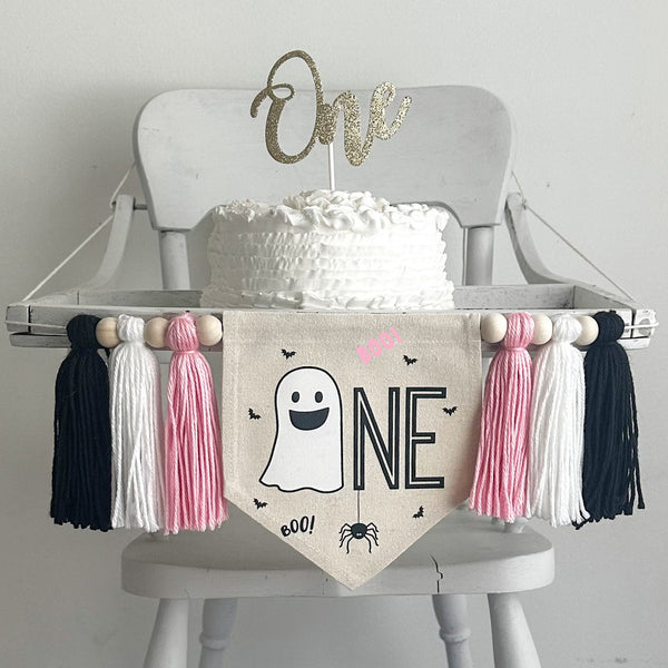 A 5 x 7 inch canvas center flag with a ghost followed by the letters NE in black and accented with a spider, bats and the word BOO in pink. On each side there is a light pink, white and black 5 inch yarn tassel, each separated by a wooden bead.