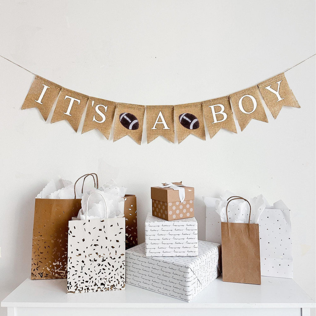 This is a burlap banner made from 4.5 by 6.5 inch fishtail shaped flags. It says IT&#39;S A BOY in white capital letters. There is a stenciled image of a football in between each word for a total of two footballs.