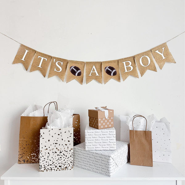 This is a burlap banner made from 4.5 by 6.5 inch fishtail shaped flags. It says IT&#39;S A BOY in white capital letters. There is a stenciled image of a football in between each word for a total of two footballs.
