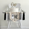 A 5x7 canvas flag with a white ghost followed by the letters NE in a black outline. There is a spider hanging from the N and bats and the word BOO all around the ONE. There is a gray, white and black tassel on each side, separated by a wooden bead.