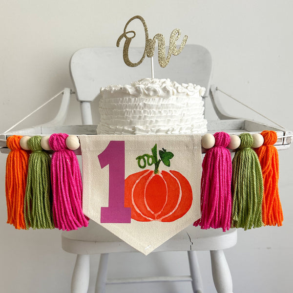 A 5 x 7 inch canvas center flag with a pink hot pink number 1 with an orange pumpkin beside it. On each side there is a hot pink, lime green, and orange 5 inch yarn tassel, each separated by a wooden bead.