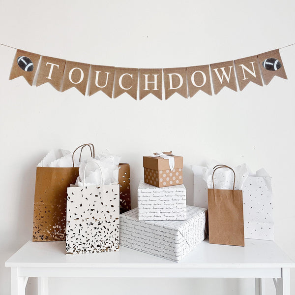 A burlap banner made from 4.5 x 6.5 inch fishtail shaped flags. There is a white stenciled letter on each flag to spell out the word TOUCHDOWN. On each end there is a hand stenciled image of a football.