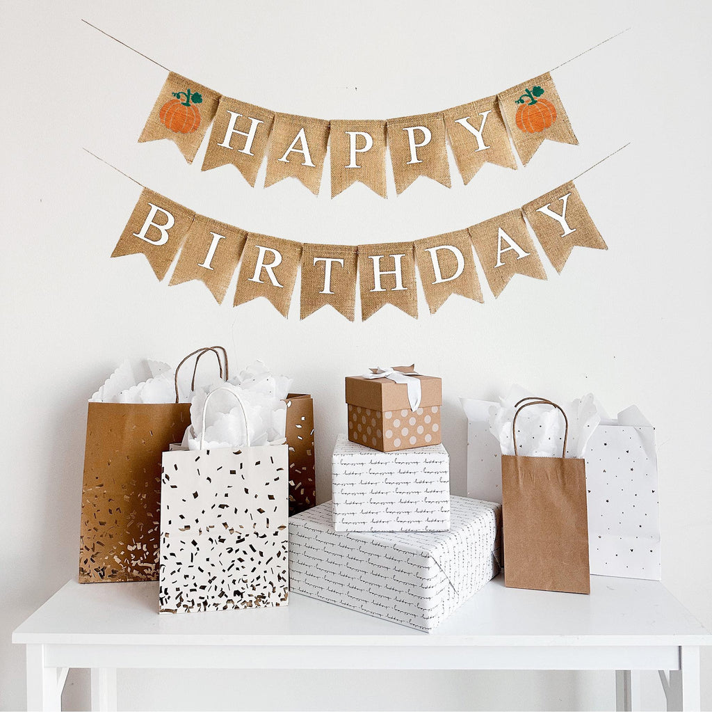 A burlap banner made from 4.5 by 6.5 inch fishtail shaped flags. The top line says HAPPY in white capital letters and has an orange pumpkin with a green stem on each end of the word. The bottom line says BIRTHDAY in white capital letters.