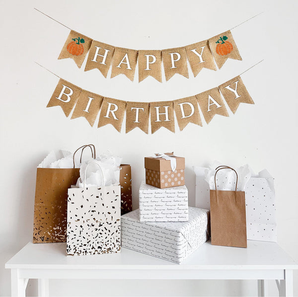 A burlap banner made from 4.5 by 6.5 inch fishtail shaped flags. The top line says HAPPY in white capital letters and has an orange pumpkin with a green stem on each end of the word. The bottom line says BIRTHDAY in white capital letters.