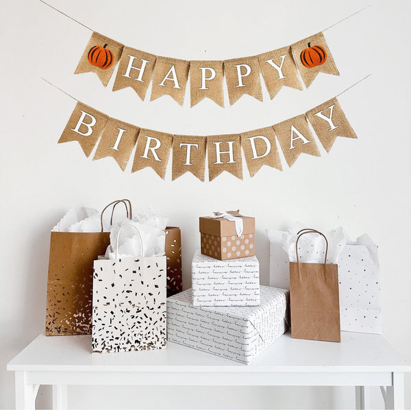 A burlap banner made from 4.5 by 6.5 inch fishtail shaped flags. The top line says HAPPY in white capital letters and has an orange and brown pumpkin with a brown stem on each end of the word. The bottom line says BIRTHDAY in white capital letters.