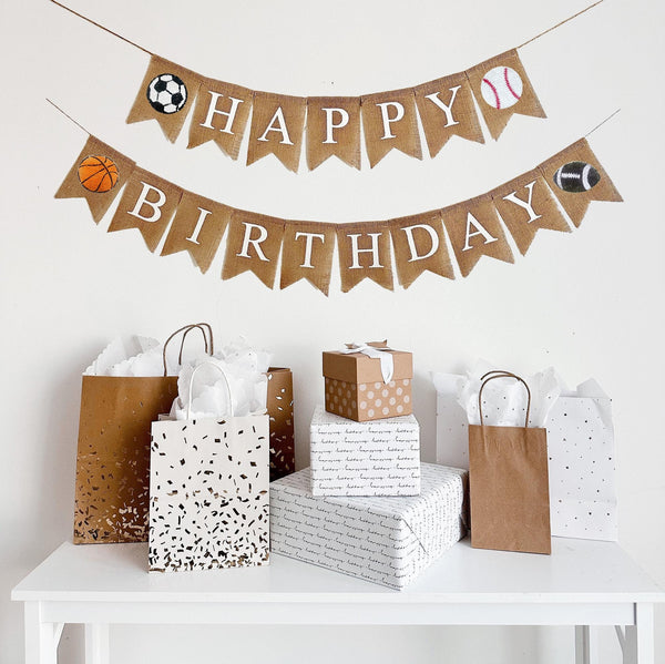 A burlap banner made from 4.5 by 6.5 inch fishtail shaped flags. The top string says HAPPY in white with a soccer ball on the left & a baseball on the right. The bottom string says BIRTHDAY with a basketball on the left & a football on the right.