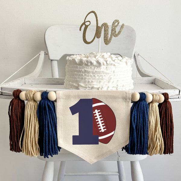 A 5 x 7 canvas flag with a large navy blue number 1 with a brown football propped against it. On each side there is a navy blue, tan and brown 5 inch yarn tassel, each separated by a wooden bead for a total of 6 tassels and 6 beads.