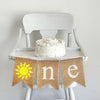 A burlap highchair banner reading one with a yellow sunshine as the o.