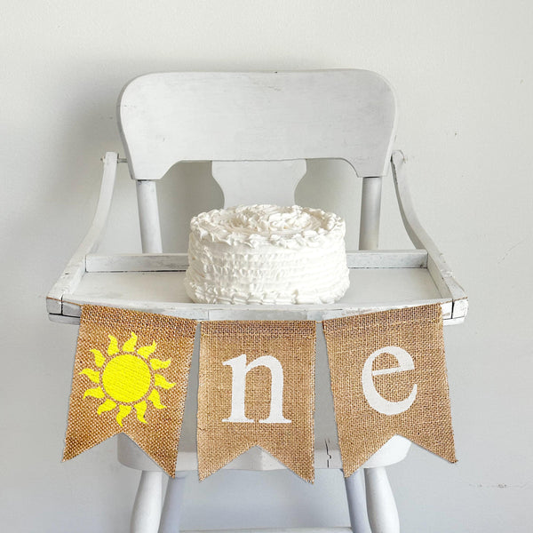 A burlap highchair banner reading one with a yellow sunshine as the o.
