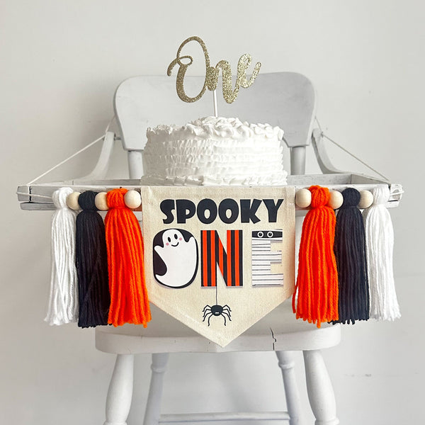 A 5 by 7 inch canvas banner with the words SPOOKY ONE and a ghost, spider and mummy. There is an orange, black and white 5 inch yarn tassel on each side, separated by wooden beads.