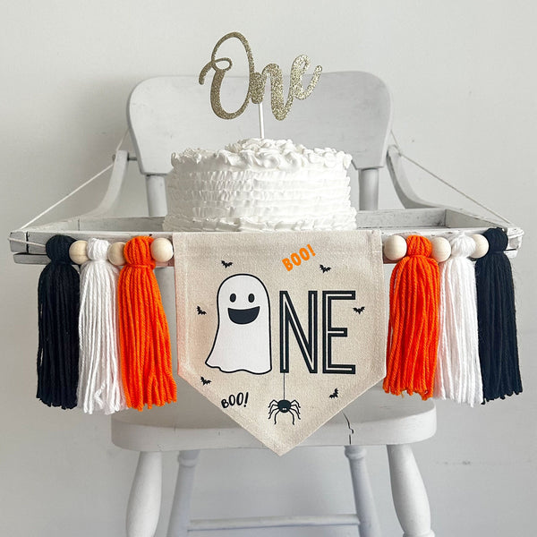 A 5 by 7 inch canvas center flag with a ghost followed by the letters NE in black, accented with a spider, bats and the word BOO. There is an orange, white and black yarn tassel on each side, all separated by wooden beads.