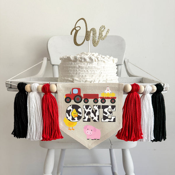 A 5 x 7 canvas banner with the word ONE in cow print. There is a red tractor above it pulling a wagon with a chicken in it. There is a duck and a pig on the flag. On each side there are red, white and black tassels, separated by wooden beads.