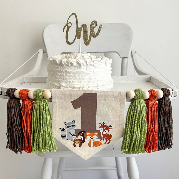 A 5 by 7 canvas banner with a large taupe number 1 surrounded by a raccoon, squirrel, fox and deer. On each side there is a 5 inch yarn sage green, dark orange and taupe tassel, each separated by a wooden bead for a total of 6 tassels and 6 beads.